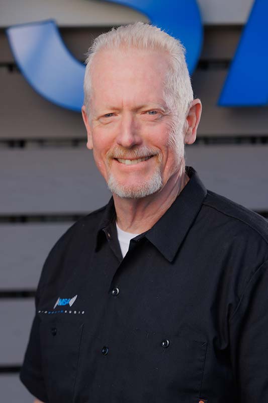 Jay-Cary-01 Smiling older man with short white hair and a goatee, wearing a black polo shirt, outdoors in front of a gray fence with blue shapes in the background.