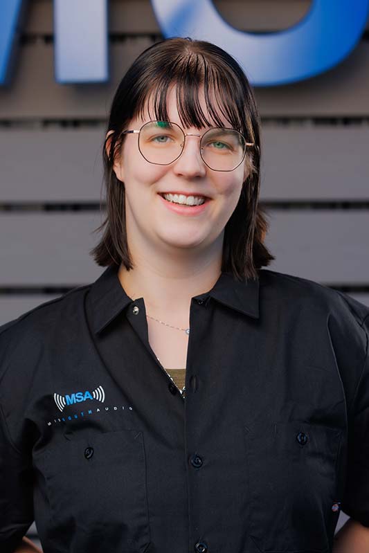 Olivia-Bangs-01 Portrait of a smiling woman with round glasses and dark hair wearing a black shirt with an MSA logo, standing in front of a gray wall backdrop.
